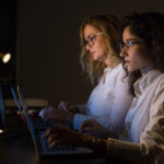 Serious businesswomen using laptops. Side view of two focused young businesswomen working with laptop computers in dark office. Working late concept